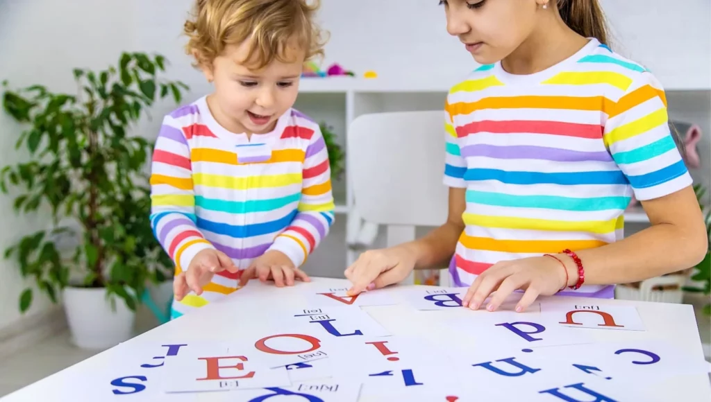 Dwoje dzieci bawiących się kolorowymi kartami z literami alfabetu - ilustracja naturalnej nauki i całkowitego zanurzenia w języku od najmłodszych lat.