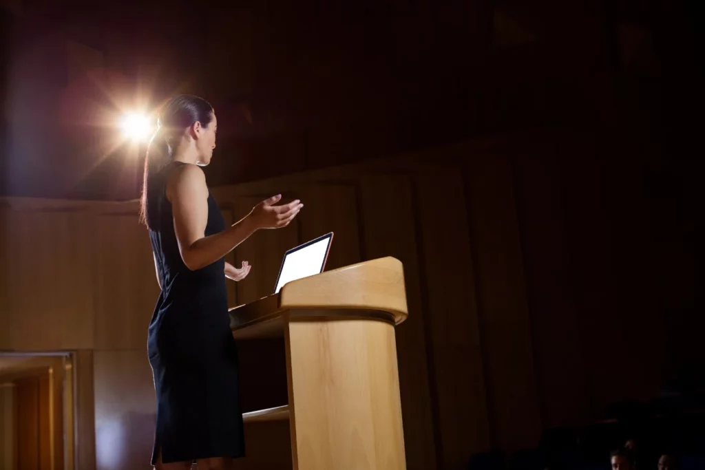Kobieta w czarnej sukience przemawiająca z podium na scenie podczas konferencji - jak wystąpienia TED inspirują do skutecznej nauki języków obcych.