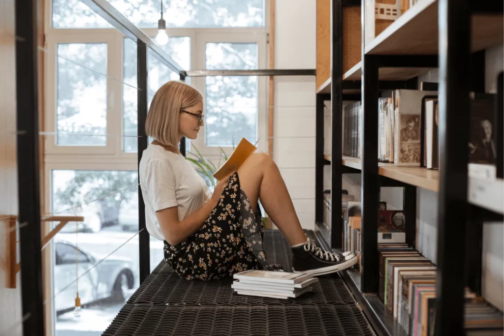 Dziewczyna w okularach czytająca książkę na antresoli biblioteki - jak wybrać najlepsze książki po angielsku dla początkujących i czerpać radość z lektury.
