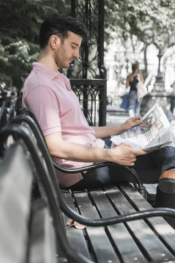 Mężczyzna w różowej koszulce czytający gazetę na ławce w parku - praktyczny przewodnik, jak efektywnie uczyć się języka z artykułów prasowych w dowolnym miejscu.