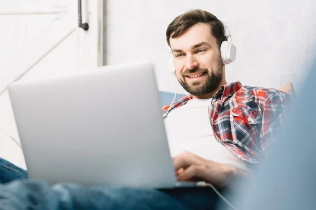 Uśmiechnięty mężczyzna z laptopem na kanapie - relaksująca i skuteczna nauka angielskiego online bez konieczności dojazdów do szkoły stacjonarnej.